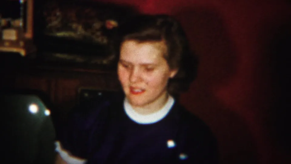 A young woman in a dark sweater and white collar smiles slightly in the 1951 Westville interior home-movie scene.