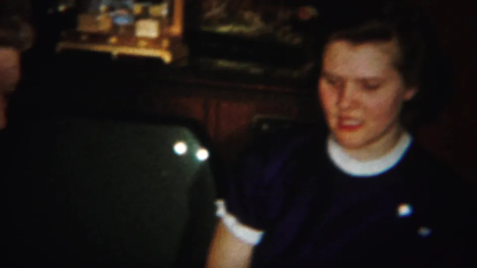 In the 1951 Westville home-movie scene, an adult woman in a dark dress and white collar sits at a card table in a dim interior.