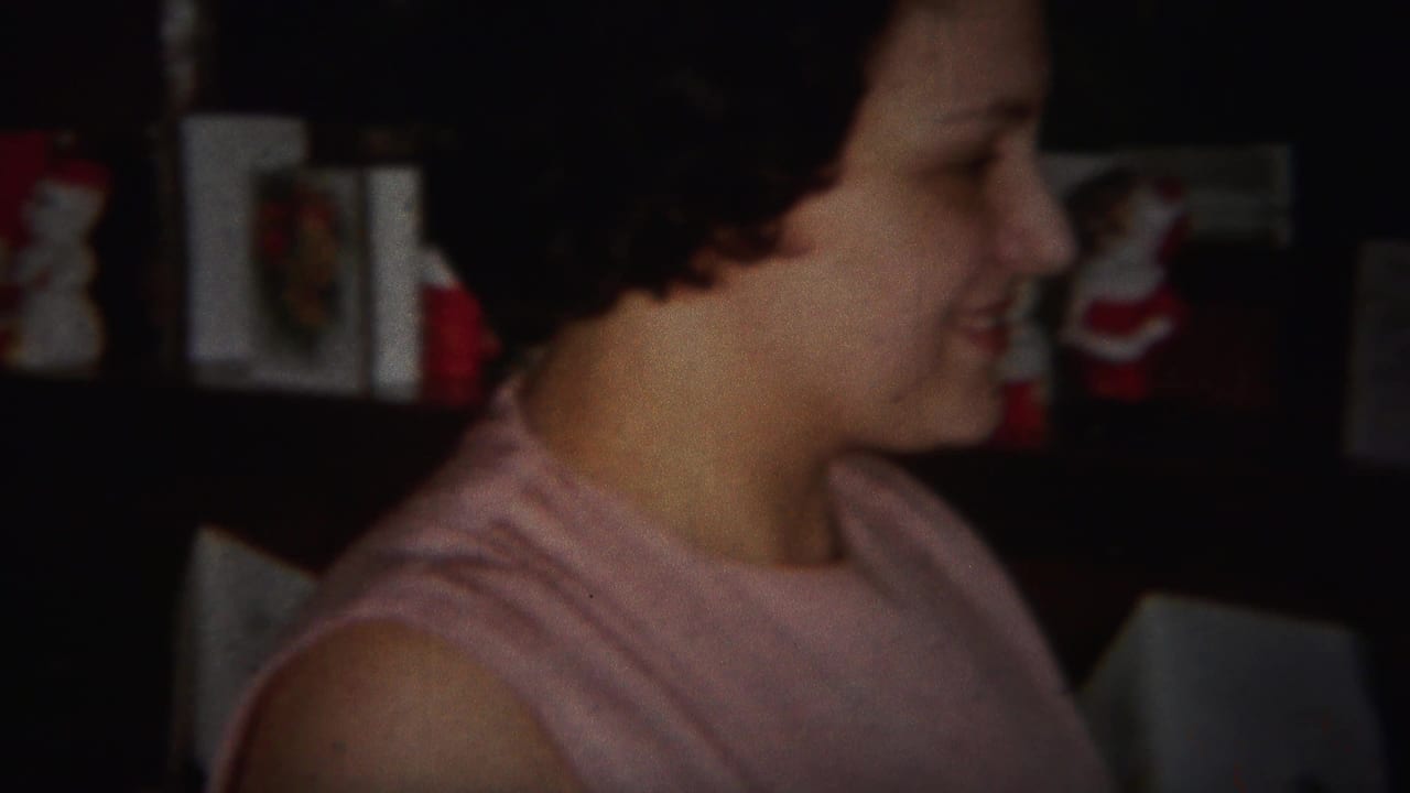 Woman in a pink sleeveless top smiles in a dim home interior with holiday figurines behind her.
