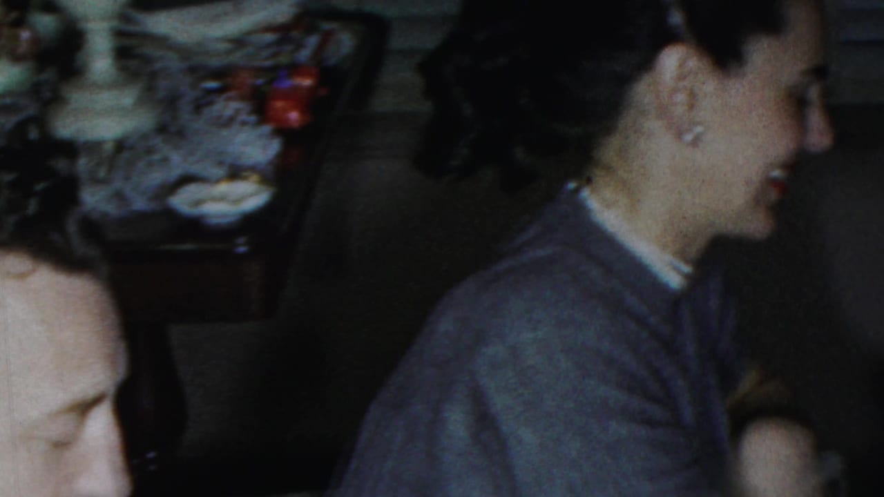 Adults seated indoors near a table, with a woman in a cardigan turned toward others.