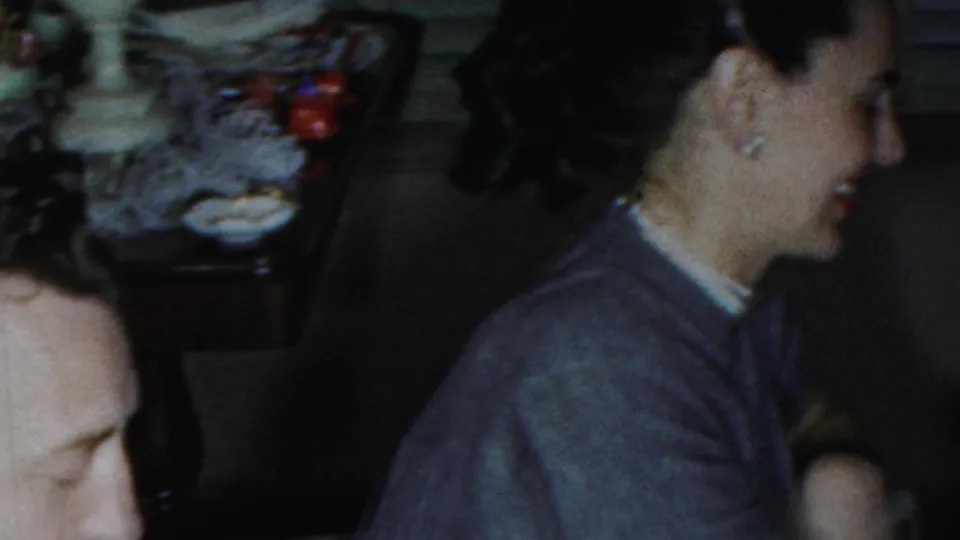 1959 Paramus home-movie frame showing adults gathered in a dim private room, with a woman in a cardigan facing the group.