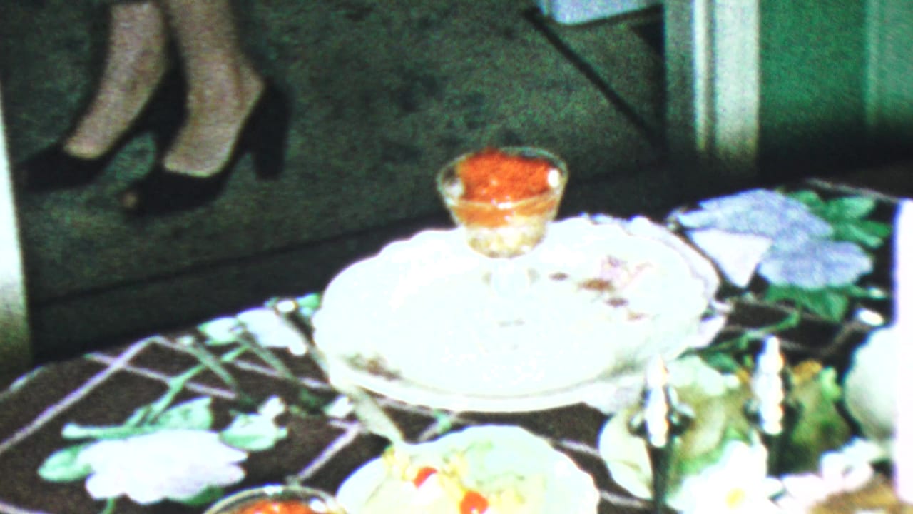 1959 Paramus home-movie frame showing a floral-covered dessert table with a white plate, glass dessert dish, and black high-heeled shoes nearby.