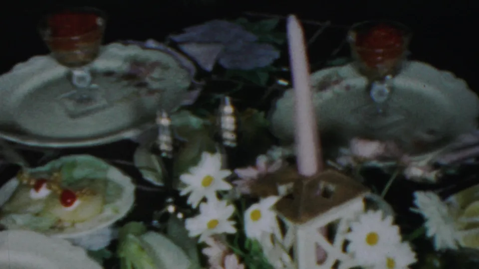 1959 Paramus home-movie frame showing a Christmas or Noche Buena table with china, stemmed red desserts, daisies, and a lantern candle.