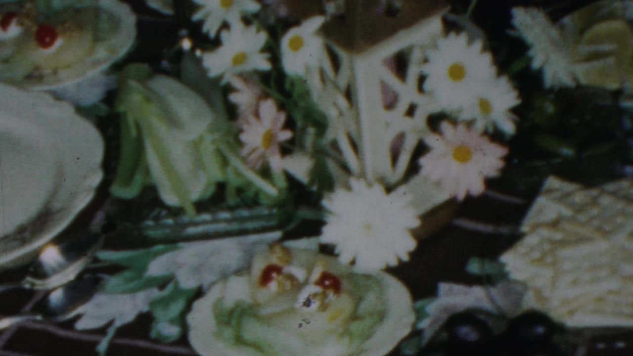 1959 Paramus home-movie frame centered on daisies, a candle lantern, white plates, pale dishes, and a dark patterned tablecloth.