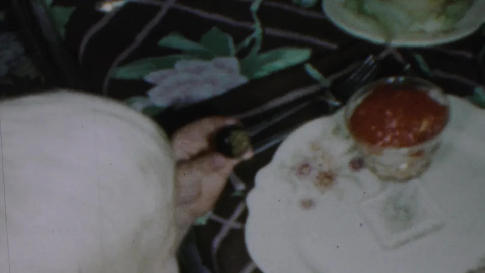 1959 Paramus home-movie frame showing floral dinner plates, pale dessert, utensils, and a small glass cup of red sauce on a patterned tablecloth.