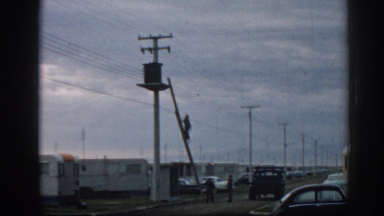 Worker on a ladder beside a utility pole with trailers, cars, and power lines under a cloudy sky.