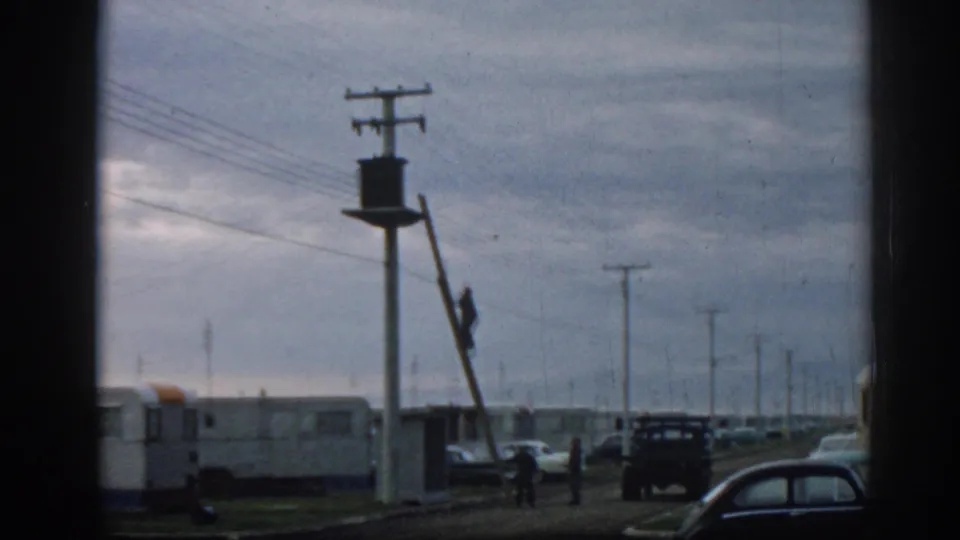 1962 Ephesus home-movie frame showing a worker climbing a ladder near a utility pole, with mobile homes, cars, and overhead wires behind him.