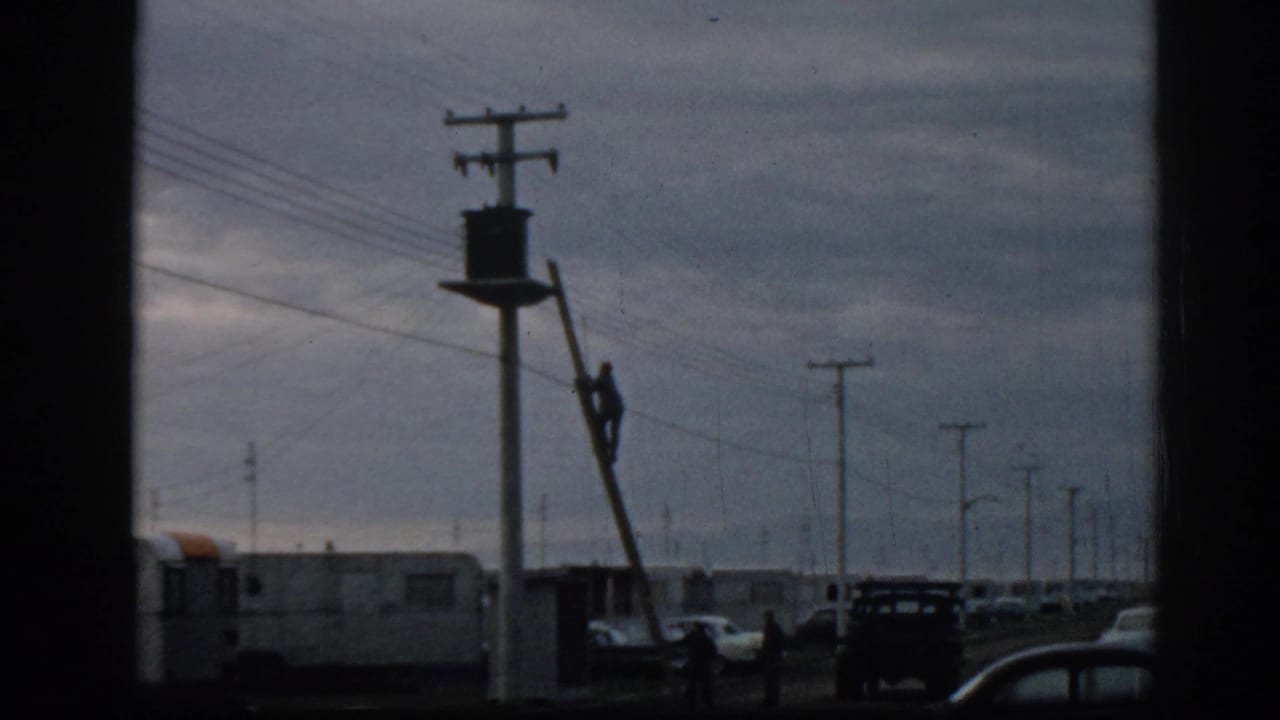 Utility worker on a ladder near a pole, with cars, trailers, power lines, and distant figures visible.