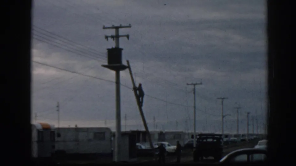 Worker repairing or inspecting a utility pole in Ephesus, Turkey, with mid-century vehicles, trailer-like homes, and power lines across the frame.