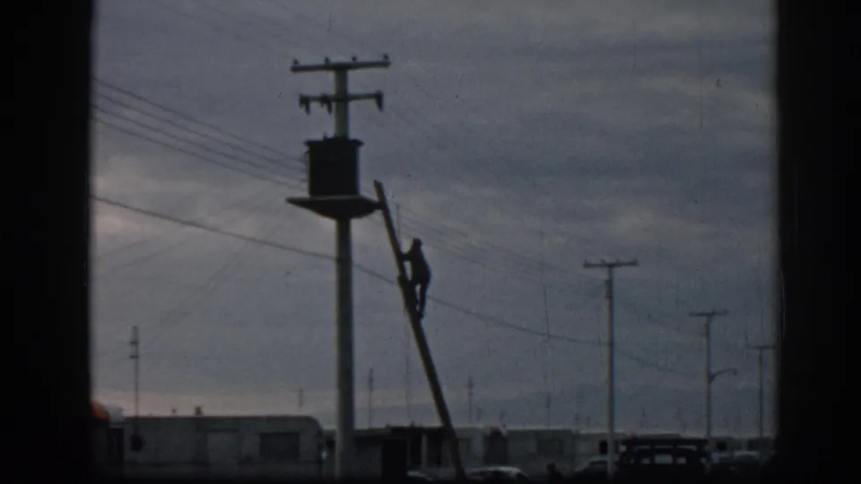 A grainy 1962 Ephesus frame shows the laddered utility worker silhouetted against clouds, with trailers, poles, wires, and cars below.