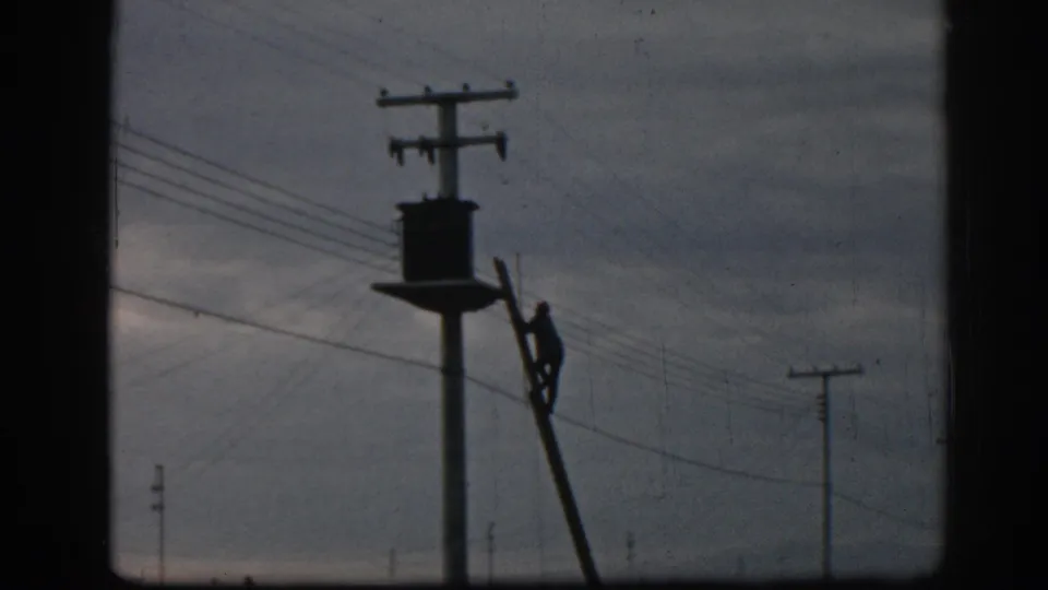 Late frame from the 1962 Ephesus clip showing the worker on a ladder near a utility pole, power lines, open sky, and roadside vehicles.