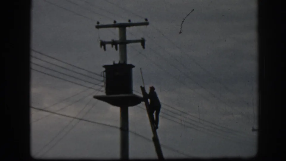 A silhouetted technician climbs beside a pole-mounted transformer with overhead lines crossing the 1962 Ephesus frame.