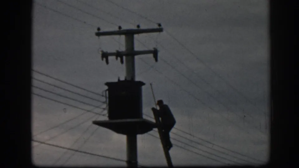 The worker is silhouetted near the transformer platform, with crossarms and multiple utility lines visible against the sky.