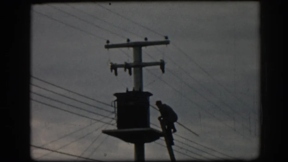 A silhouetted utility worker remains on the ladder beside a pole-mounted transformer in archival 8mm footage from Ephesus, Turkey.