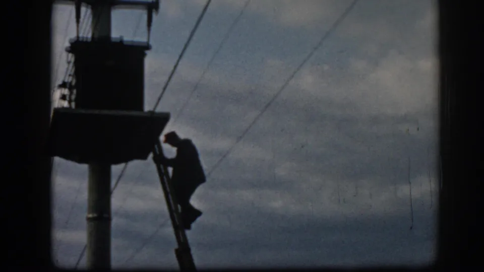 In 1962 Ephesus footage, a silhouetted technician is partway down a ladder beside a utility pole under a cloudy sky.