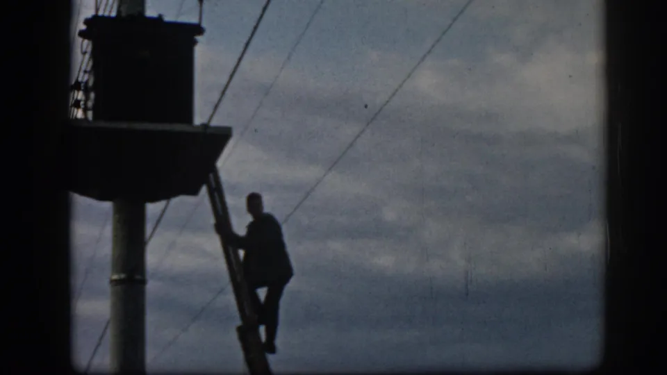 The worker appears in silhouette on the ladder beside a pole and overhead lines in the 1962 Ephesus home-movie frame.