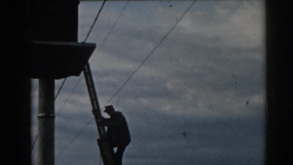 A silhouetted technician is lower on the ladder below utility equipment, with wires crossing the cloudy sky in Ephesus, Turkey.
