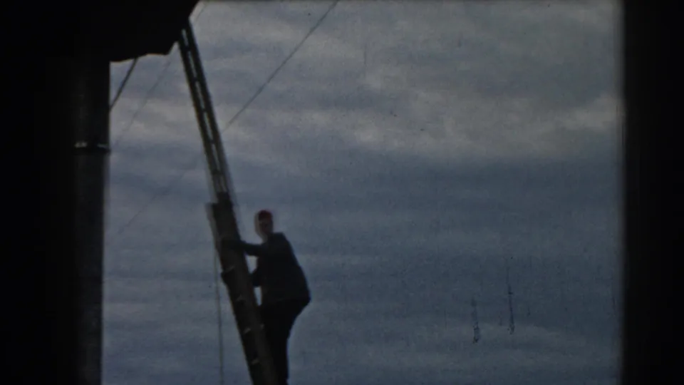 In 1962 Ephesus, Turkey, a worker in dark clothing and a red cap climbs a ladder near overhead wires under a cloudy sky.