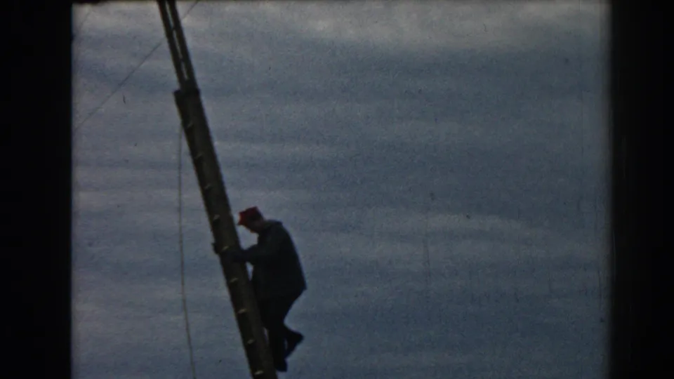 The worker in dark clothing leans into the ladder in this 1962 Ephesus home-movie frame, with wires and cloudy sky behind him.