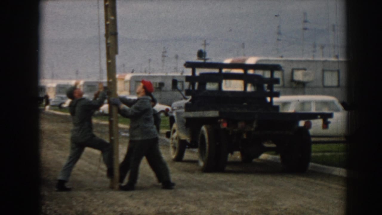 In 1962 Ephesus, Turkey footage, three men brace a tall wooden pole beside a dark flatbed truck with trailers and cars behind them.