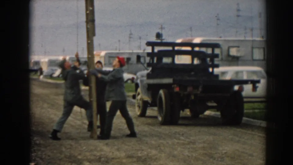 The men continue bracing the pole in a trailer yard, with one wearing a red cap and a dark flatbed truck parked nearby.
