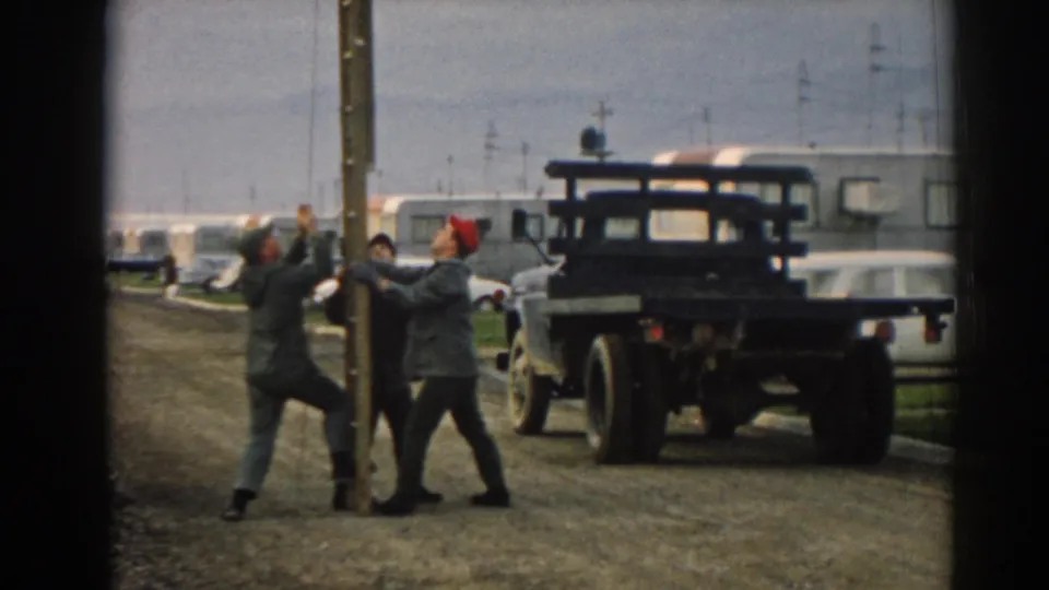 Three men work close to the pole base in the 1962 trailer yard scene, framed by a dark truck, trailers, and older cars.