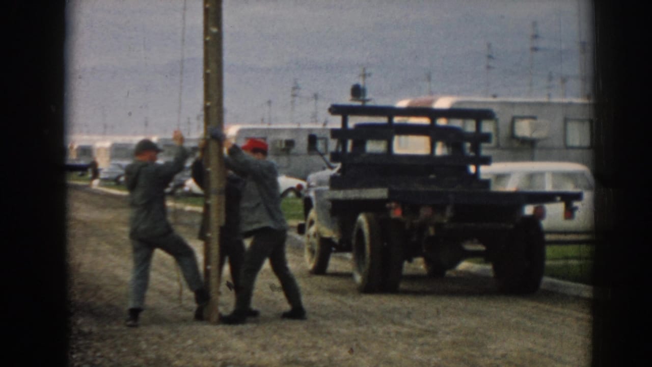 Three men steady a large wooden pole beside a parked flatbed truck on a roadside.