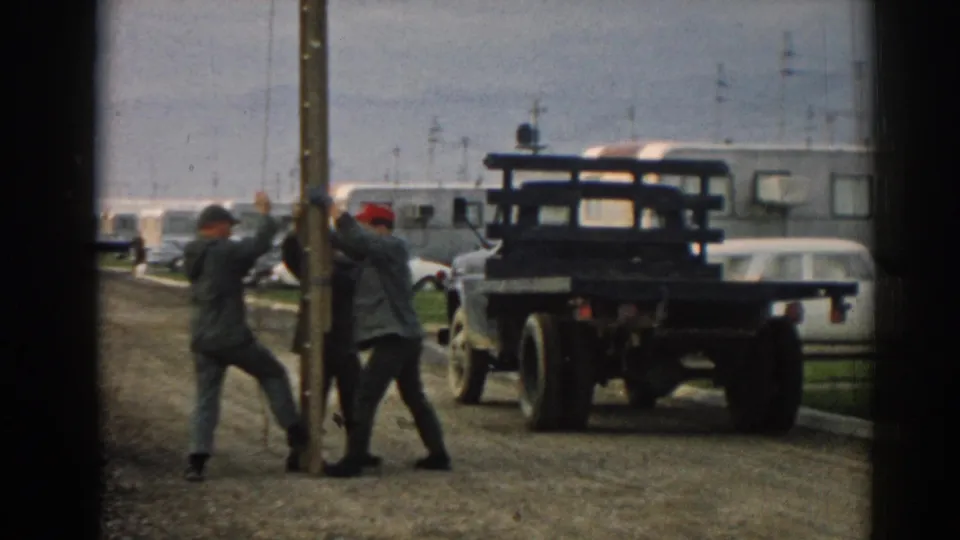 In 1962 Ephesus, Turkey, three adult men appear to push and steady a large wooden utility pole near a dark flatbed truck and parked cars.