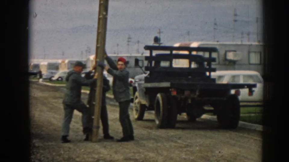 The tall wooden pole is nearly upright as two men hold it and a third man stands nearby, with a vintage flatbed truck and cars in the background.