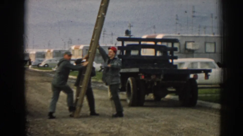 At the end of the clip, the men brace the leaning wooden pole beside the road, with the flatbed truck, parked cars, and trailer-like structures behind them.