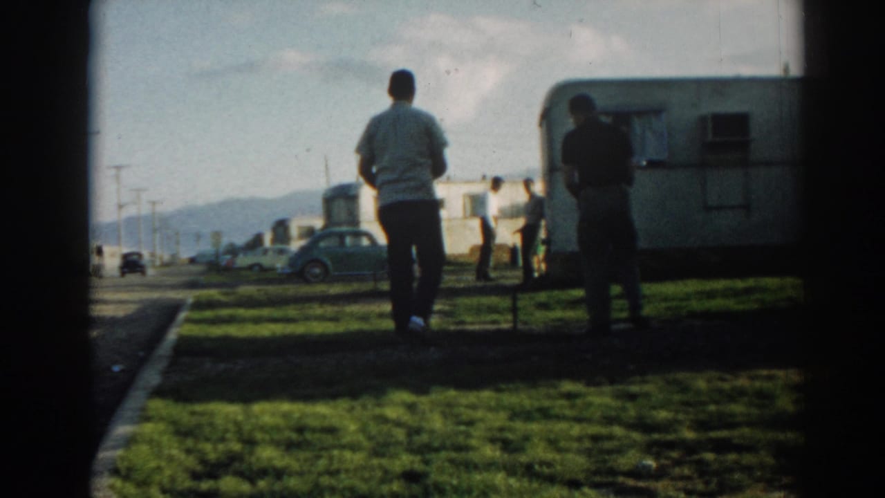 Two men stand on grass near a camper trailer, with parked cars and hills in the background.