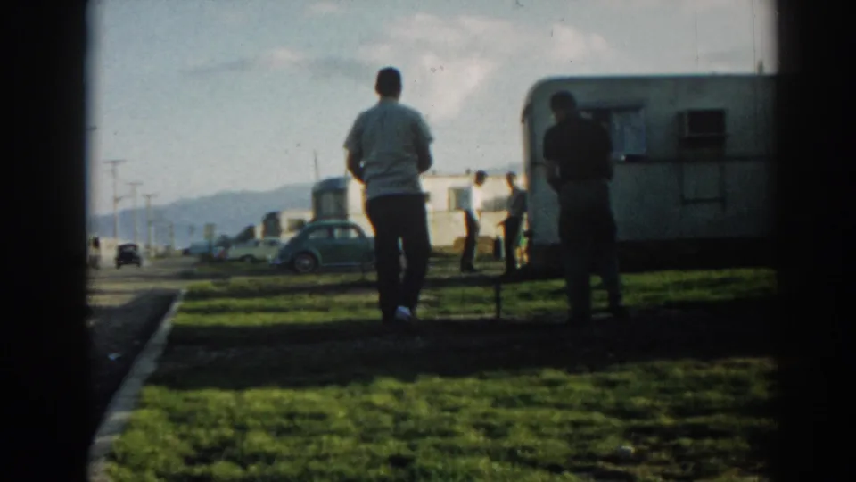 1962 Ephesus, Turkey home-movie frame showing two adult men at a grassy camper site with parked cars, a white trailer, and distant hills.