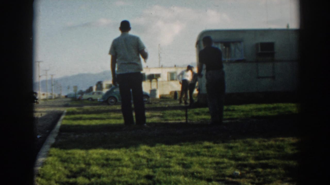 Men stand near a camper trailer and parked cars on a sunny grassy campsite.