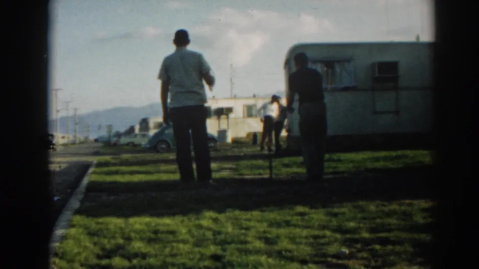 Adult men gather near a white camper trailer at the Ephesus campsite, with a small car and hills visible beyond the grass.
