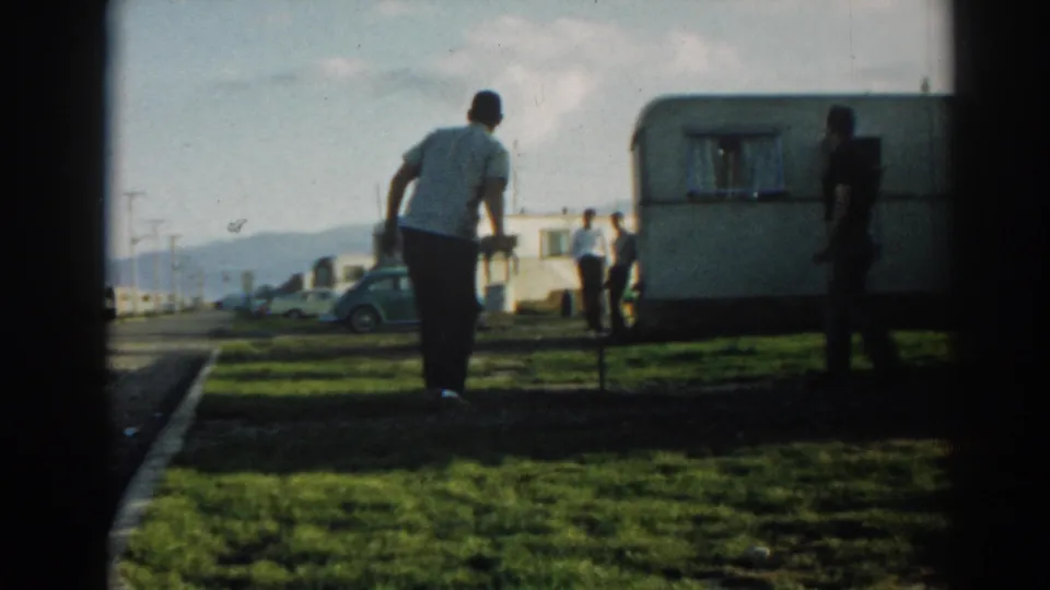A man bends toward the horseshoe area at the Ephesus camper site, with another man, a trailer, parked cars, and hills in view.