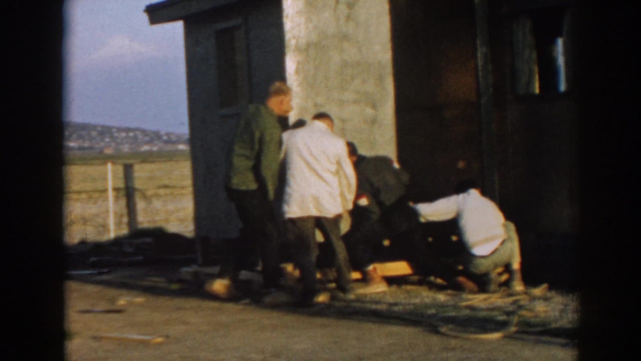 Several men in 1960s clothing gather by a small building in Ephesus, Turkey, with wood pieces and rope or cable at their feet.
