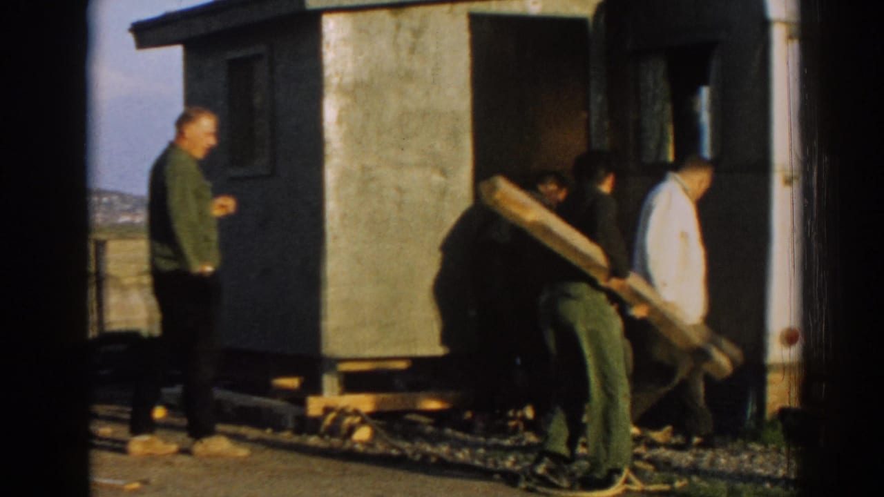 Men handle a long wooden plank beside a small building in 1962 Ephesus, with another man standing nearby in the open setting.