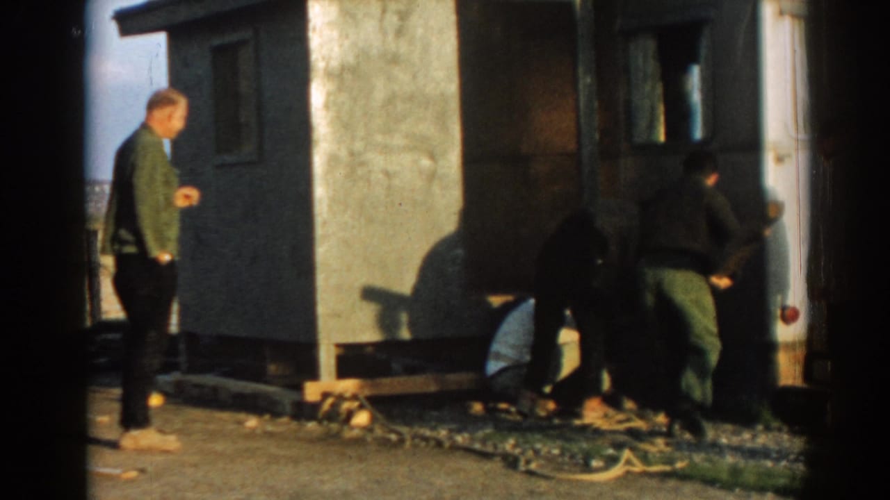 Men stand and bend beside a small structure while another man watches from the left.