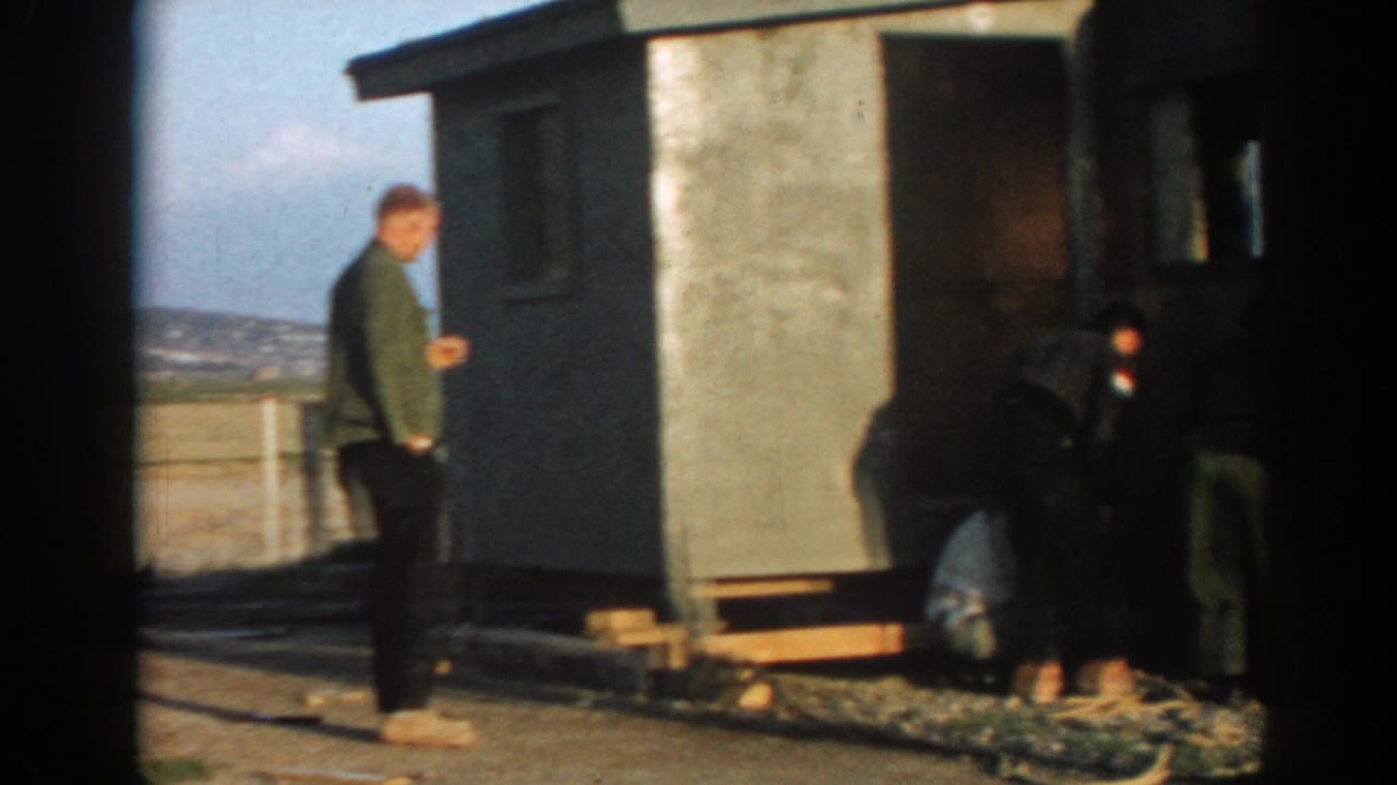 A man in a green jacket stands near a small prefabricated structure while other men work low to the ground in Ephesus, Turkey.