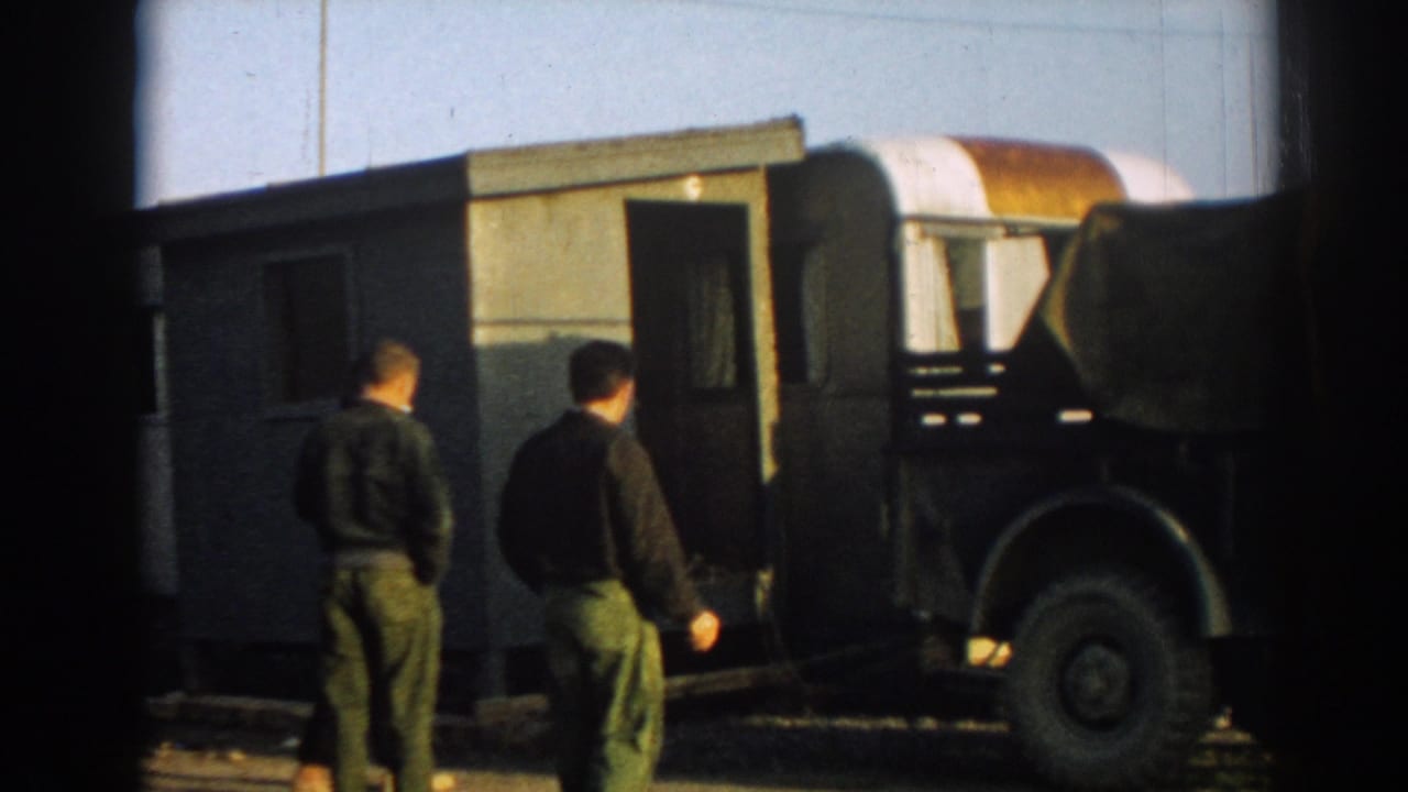 Two men in military-style clothing stand beside a dark green truck and towable trailer in 1962 Ephesus, Turkey footage.