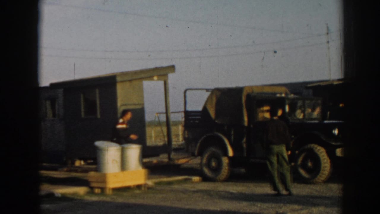 People stand near a green wooden structure, metal drums, and a canvas-topped jeep.