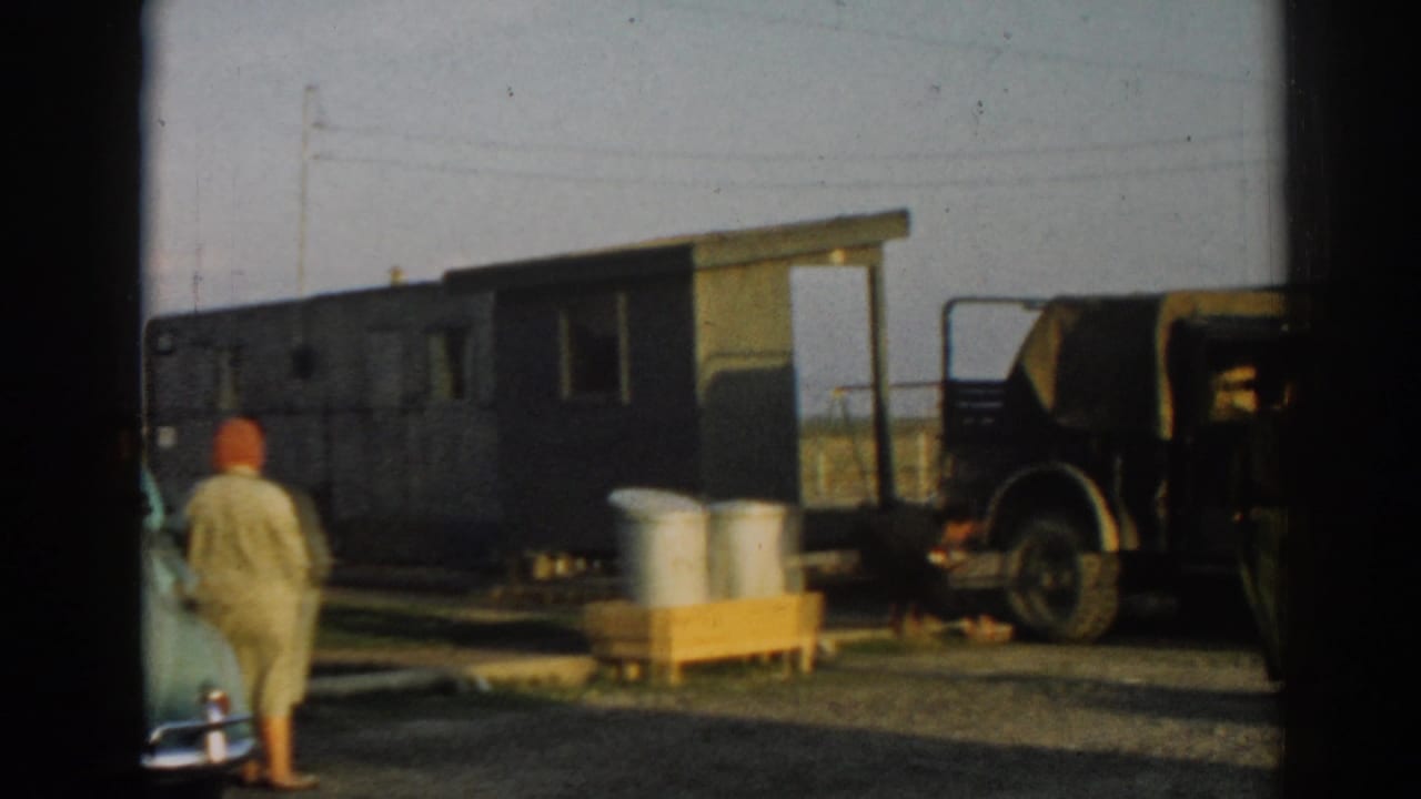 1962 Ephesus, Turkey frame with a person walking near a green wooden structure, metal drums, a jeep, and open ground.