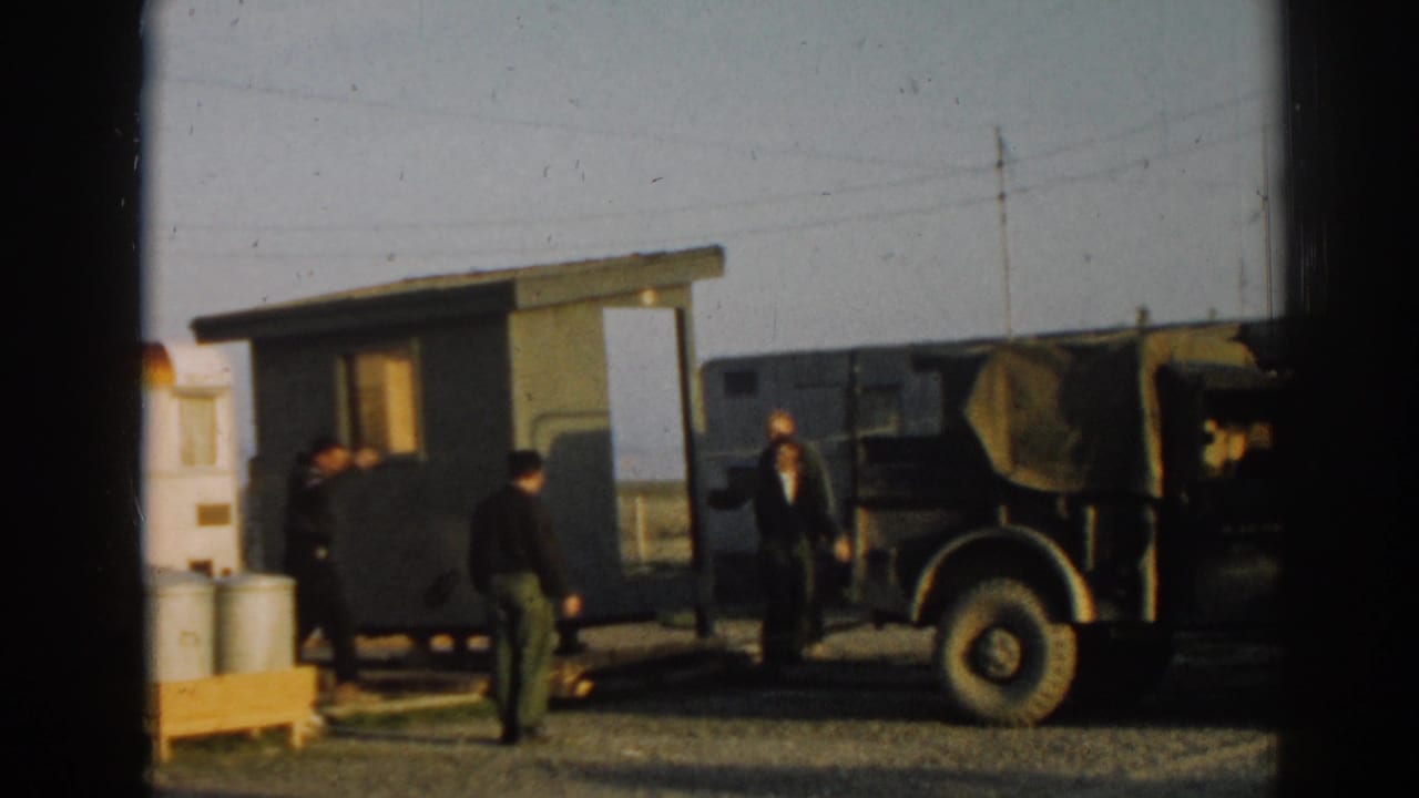 1962 Ephesus, Turkey frame with people around a green wooden structure, a canvas-topped jeep, metal drums, and a partial green car.