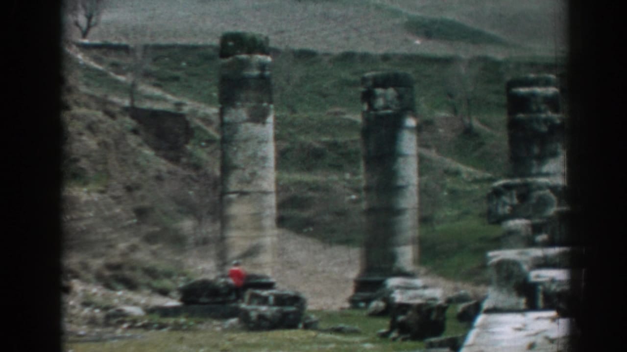 Grey stone columns and ruins at the base of hills with a small red-clad person nearby.