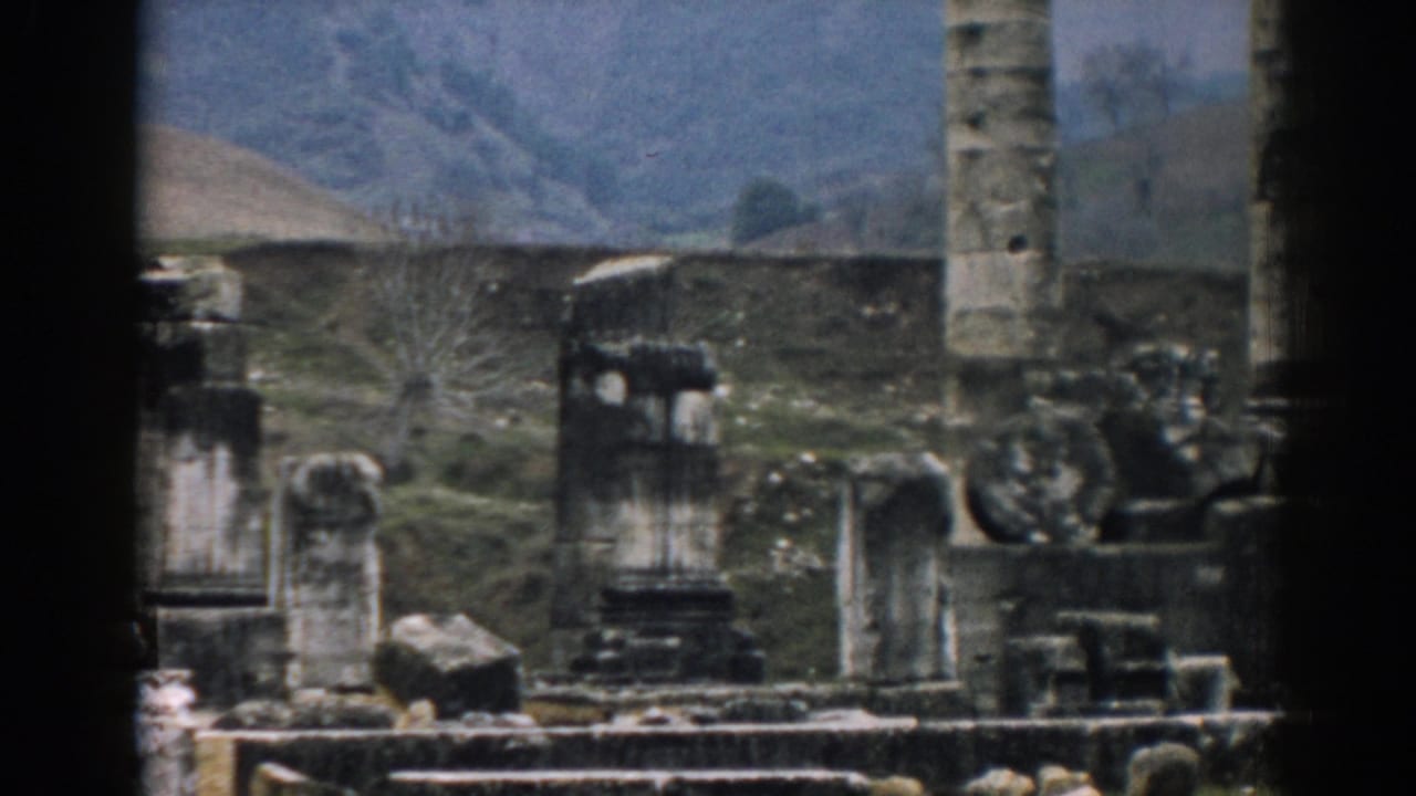 Handheld 8mm frame from Ephesus with a tall column, weathered ruin fragments, low stonework, and open terrain.