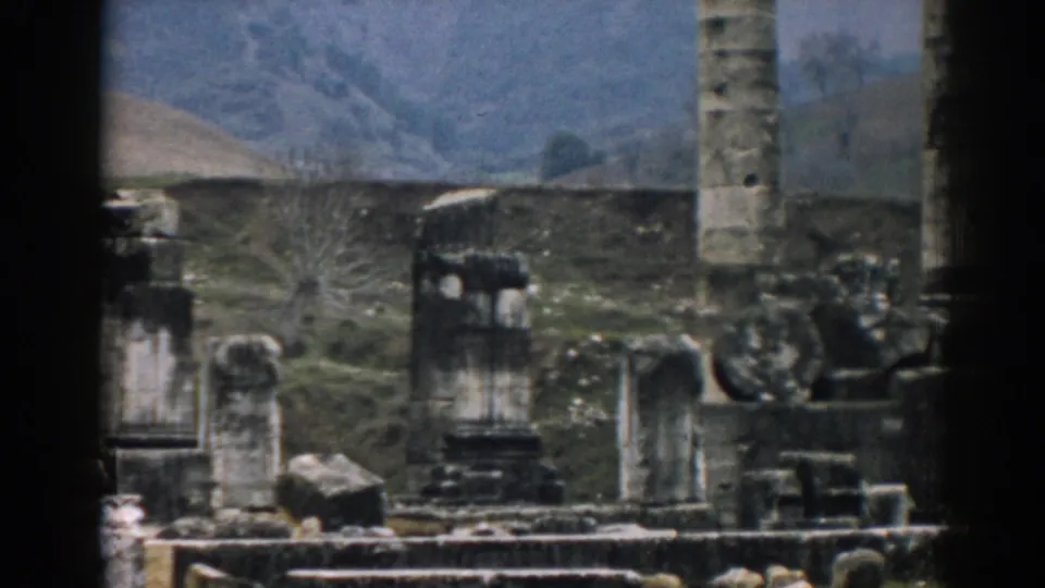 Handheld 8mm frame from Ephesus with a tall column, weathered ruin fragments, low stonework, and open terrain.