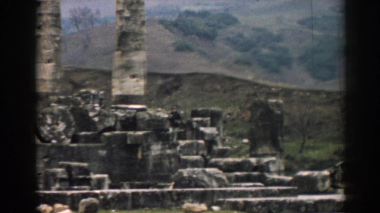 1962 Ephesus travel footage frame with a distant visitor among broken stonework beneath a tall column and hills.