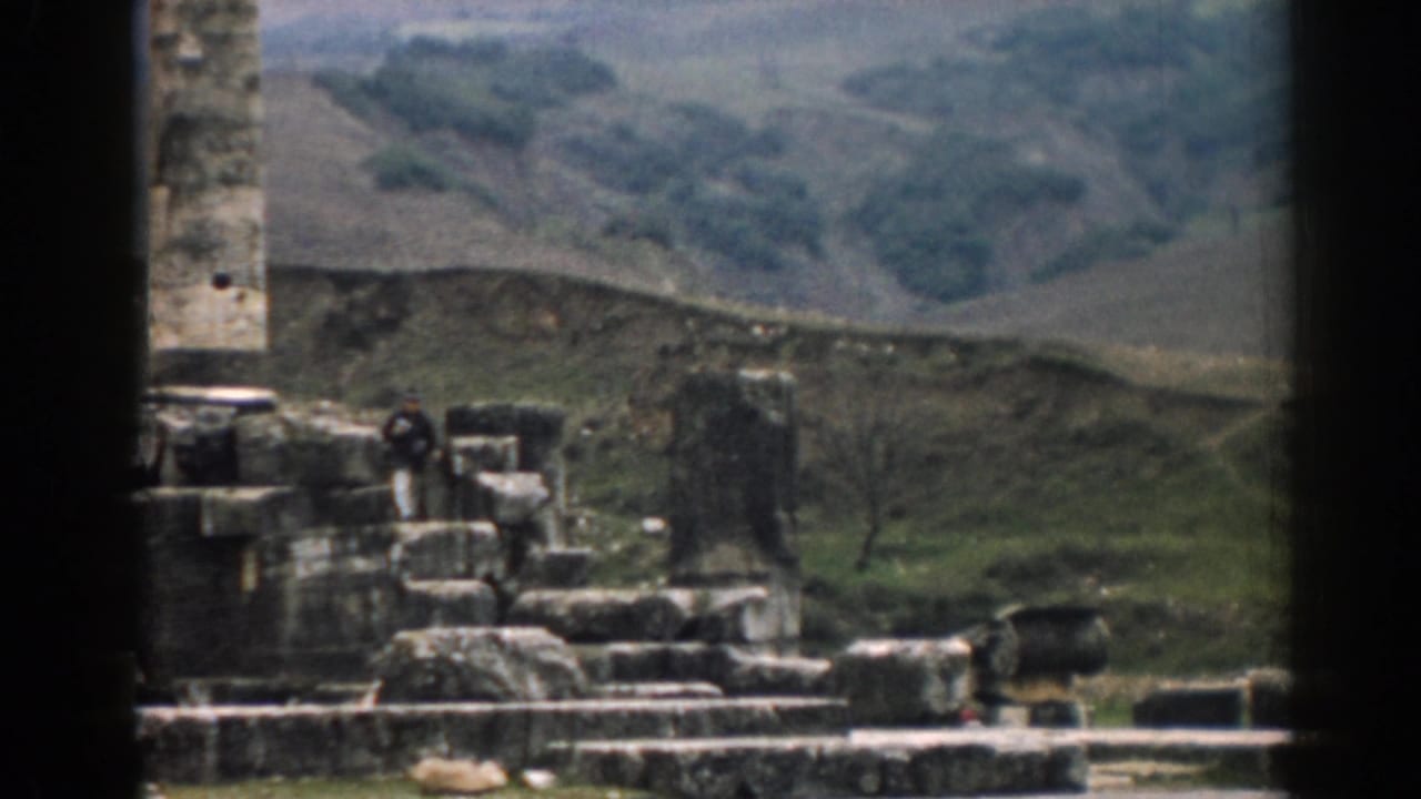 1962 8mm frame of Ephesus ruins with a distant dark-clothed visitor, stone fragments, and rolling hills.