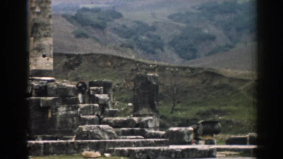 1962 8mm frame of Ephesus ruins with a distant dark-clothed visitor, stone fragments, and rolling hills.