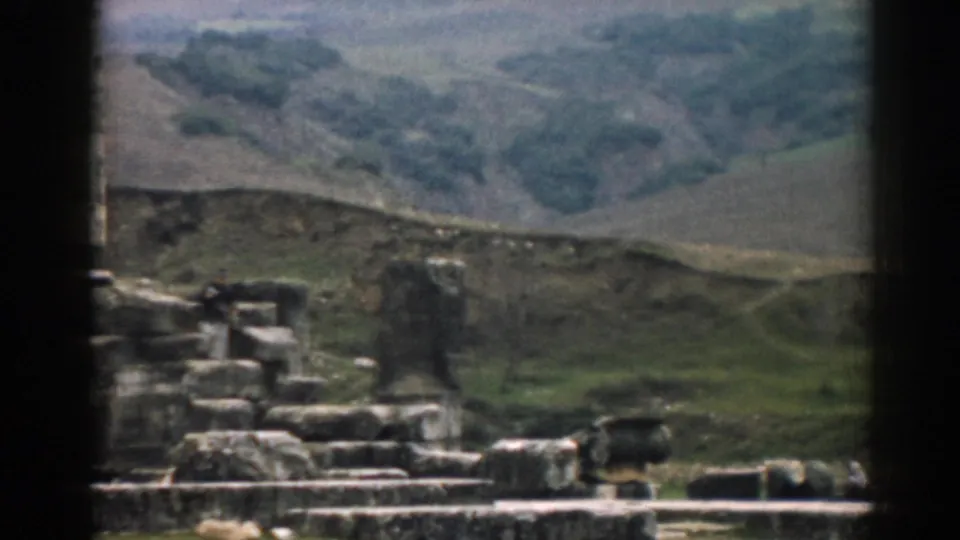 1962 Ephesus home-movie frame showing broken stonework, a stepped ruin, and an adult visitor against rolling hills.